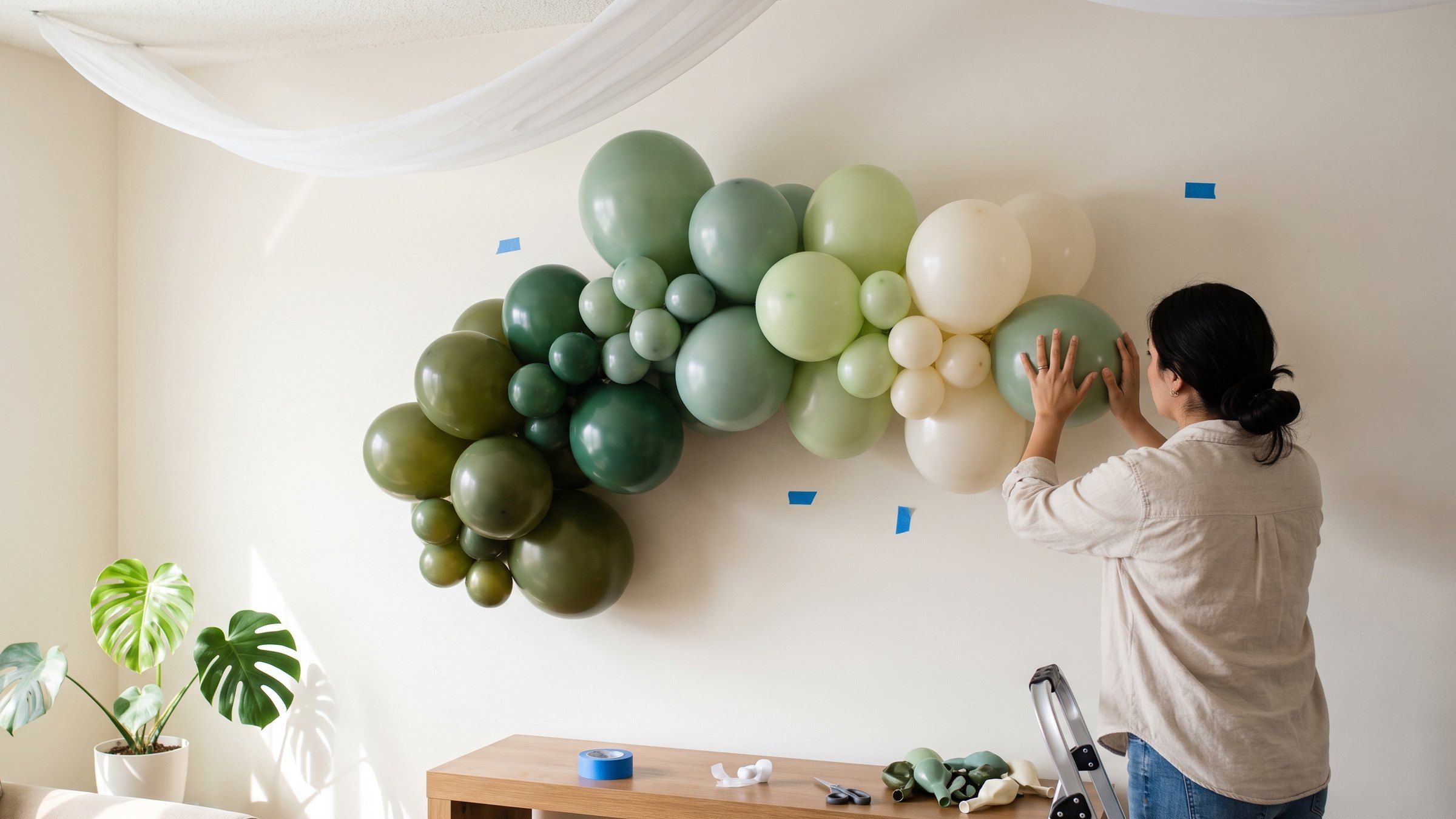 Mujer pega un globo verde en la pared de una sala, sobre una nube de globos en degradé, con tela blanca drapeada en el techo