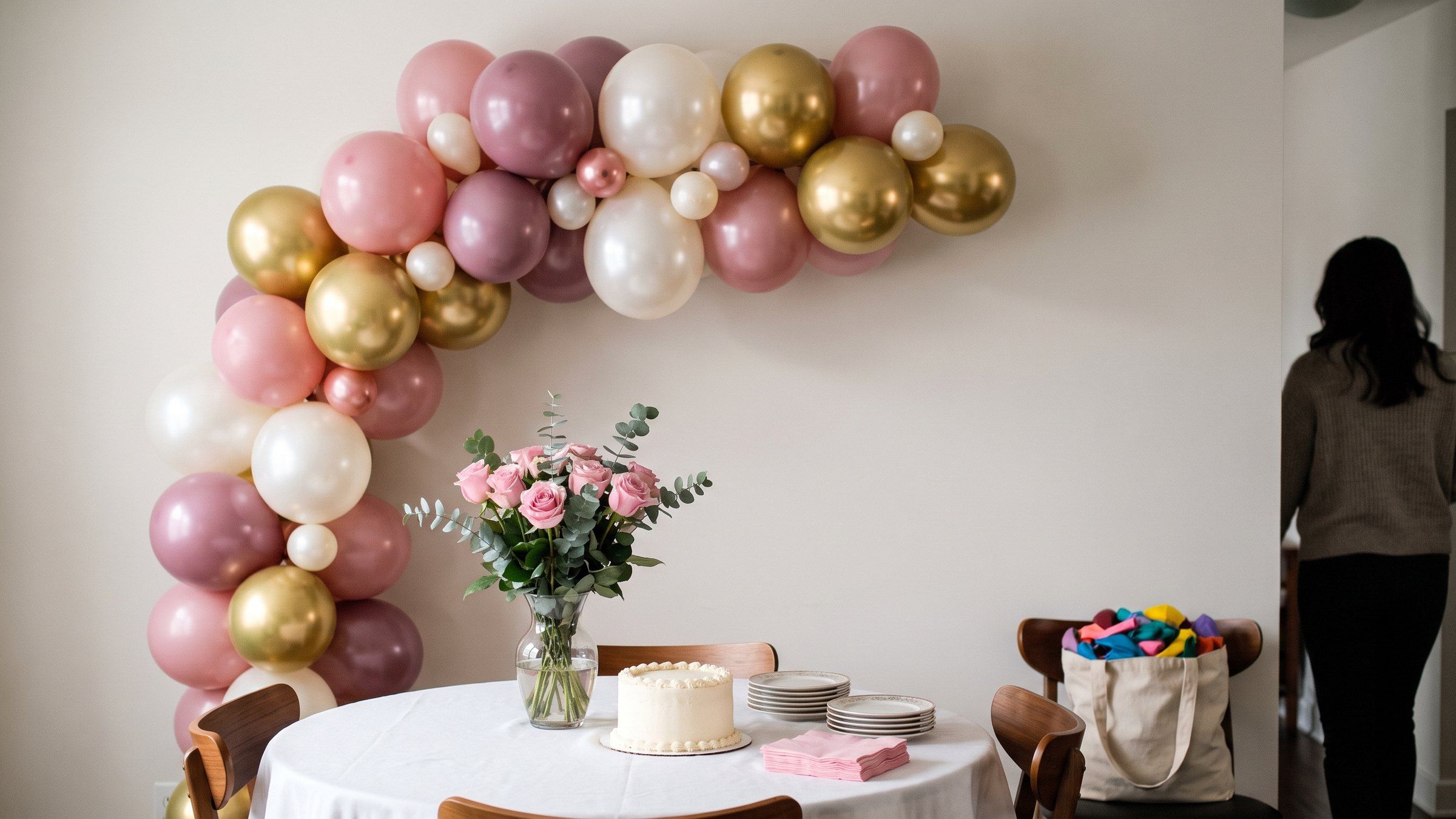 Guirnalda de globos rosa empolvado, malva, blanco perla y dorado cromado en la pared, sobre una mesa con pastel blanco, rosas y servilletas rosadas